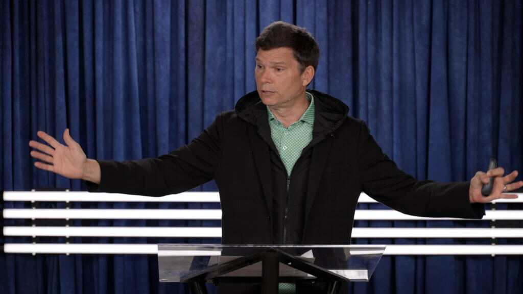 Man behind a clear lectern with arms outstretched, speaking to an audience against a blue curtain backdrop on stage for a talk or sermon.